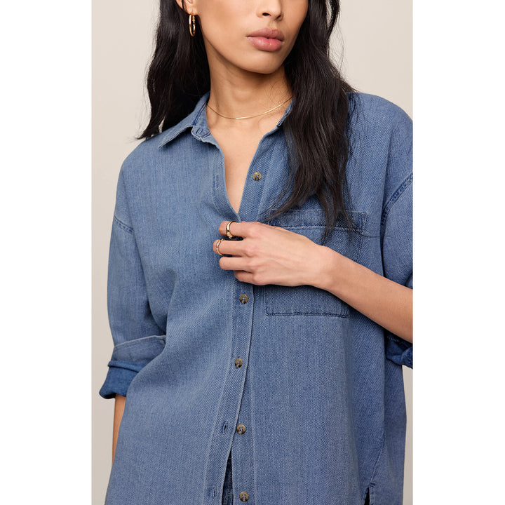 A woman wearing an oversized blue denim button-up shirt, shown from the chest down against a plain light beige background.