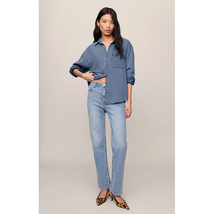 A model stands against a plain beige studio background wearing a cropped blue denim button-up shirt, light-wash blue jeans, and leopard-print heels.