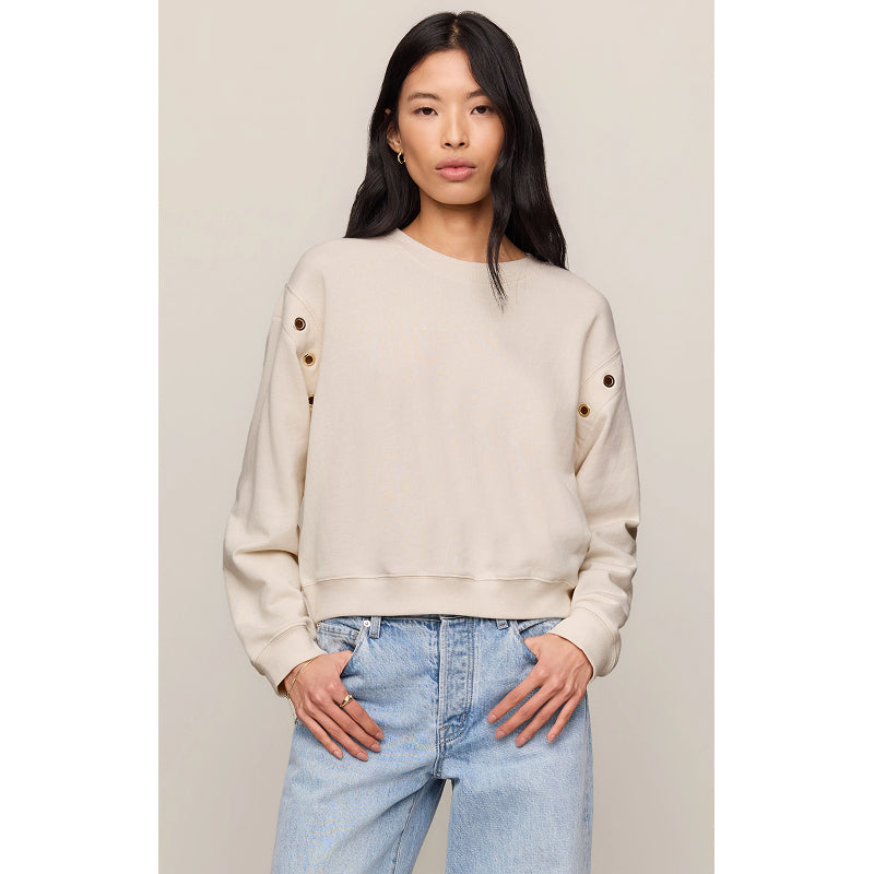 A woman modeling a cropped cream-colored sweatshirt with decorative grommets on the sleeves, paired with light-wash blue jeans against a neutral beige background.