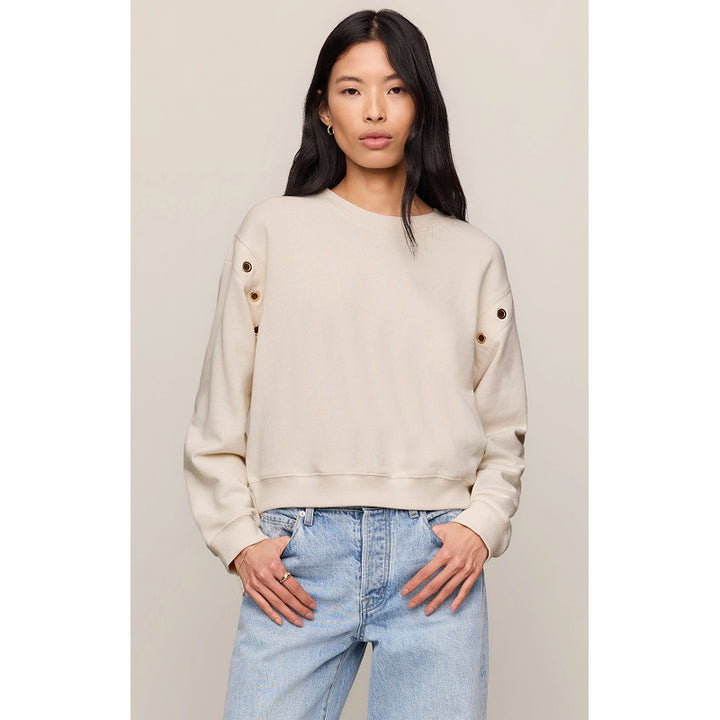 A woman modeling a cropped cream-colored sweatshirt with decorative grommets on the sleeves, paired with light-wash blue jeans against a neutral beige background.