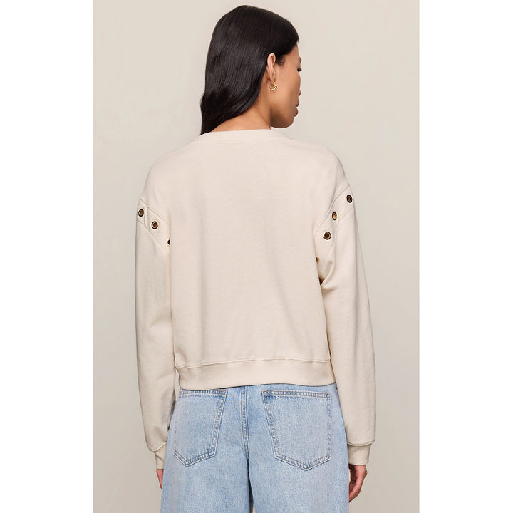 A woman shown from the back wearing a cream-colored cropped sweatshirt with decorative grommets on the sleeves, paired with light blue jeans, against a solid light beige background.