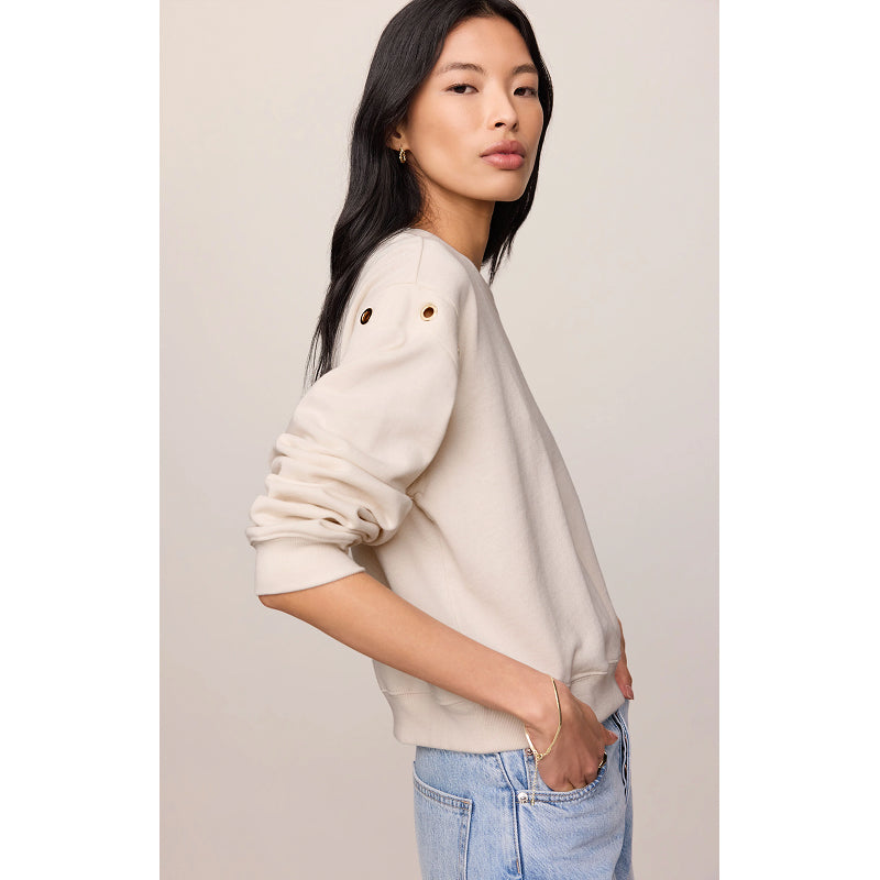 A woman wearing a cream-colored crewneck sweatshirt with decorative gold grommets on the upper sleeve, paired with light blue jeans, set against a plain beige studio background.