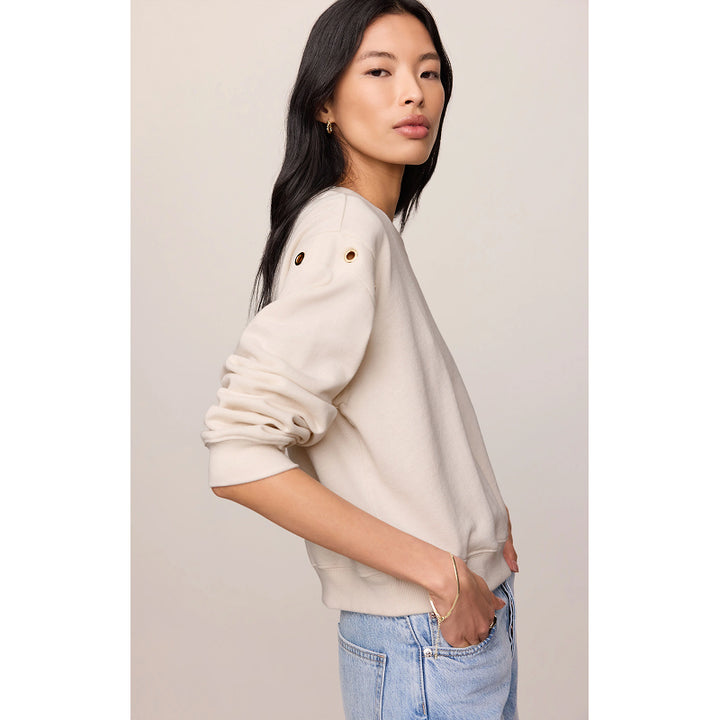 A woman wearing a cream-colored crewneck sweatshirt with decorative gold grommets on the upper sleeve, paired with light blue jeans, set against a plain beige studio background.