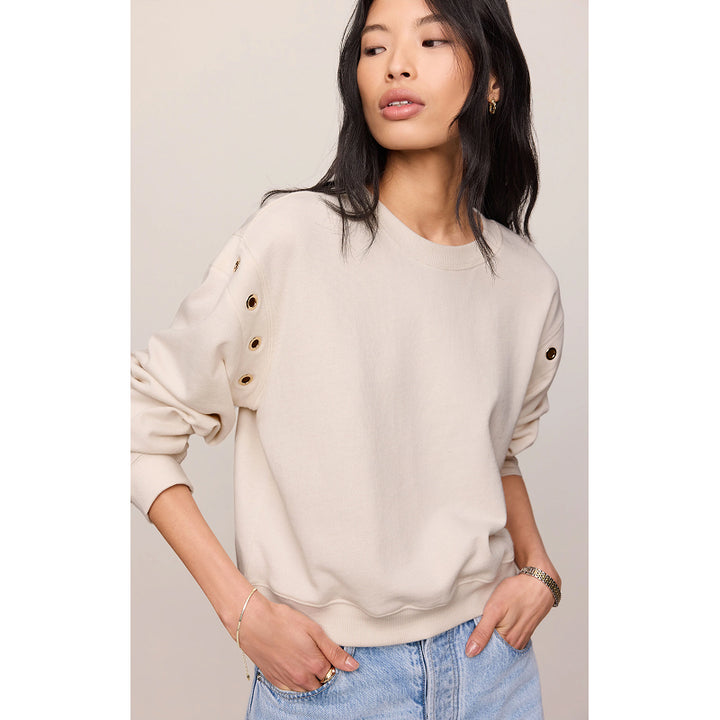 A woman wearing a light beige crewneck sweatshirt with decorative gold-tone grommet details on the sleeves, paired with light blue jeans, set against a plain light beige background.