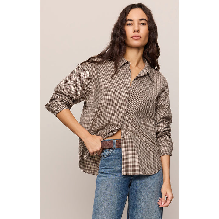 A woman modeling an oversized brown and beige gingham button-down shirt with rolled-up sleeves, paired with blue jeans and a brown belt, against a solid light beige background.