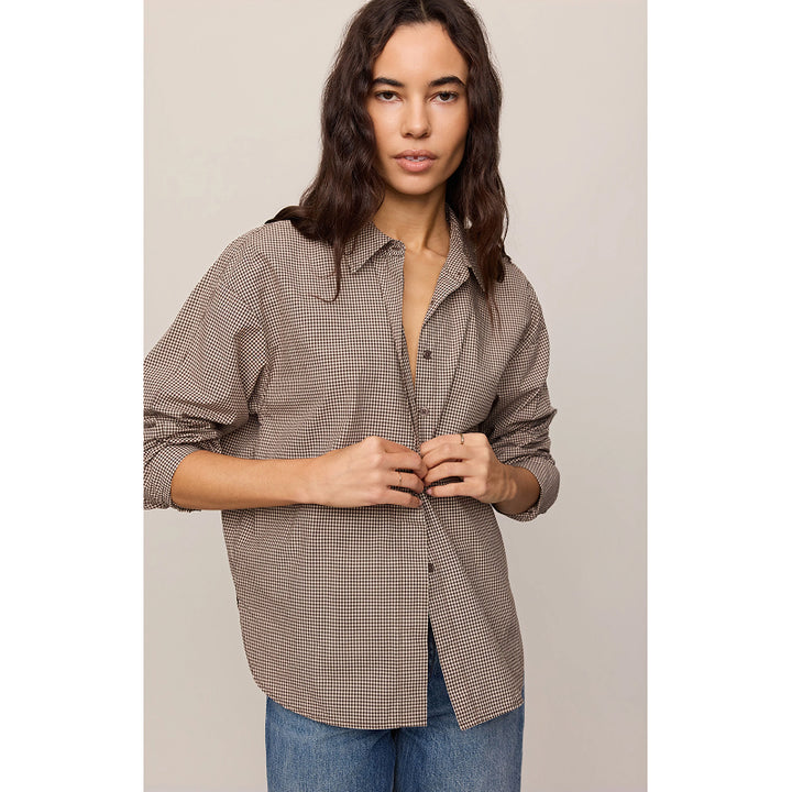 A woman wearing a brown and beige gingham check long-sleeve button-up shirt with blue jeans, standing against a neutral beige background.
