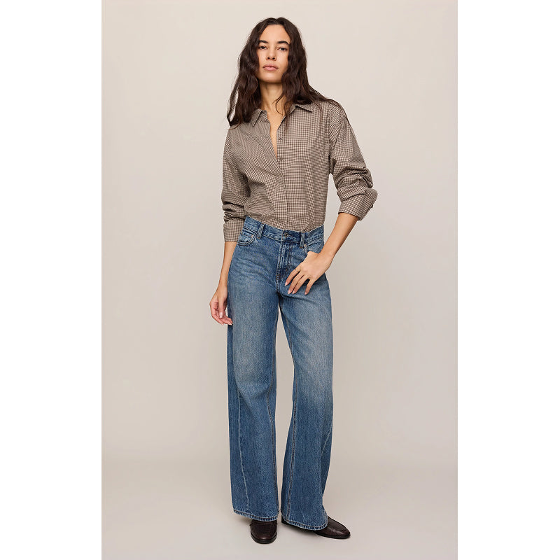 A woman modeling a brown and beige checkered button-down shirt with rolled-up sleeves and high-waisted, wide-leg blue jeans, standing against a neutral beige studio background.