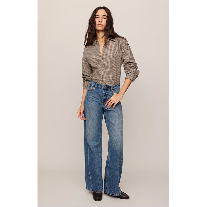 A woman modeling a brown and beige checkered button-down shirt with rolled-up sleeves and high-waisted, wide-leg blue jeans, standing against a neutral beige studio background.