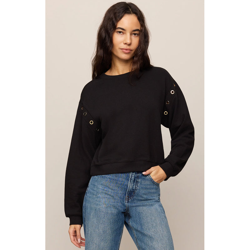 A woman modeling a black long-sleeve crewneck sweatshirt with gold grommet details on the sleeves, paired with blue jeans, against a solid light beige background.