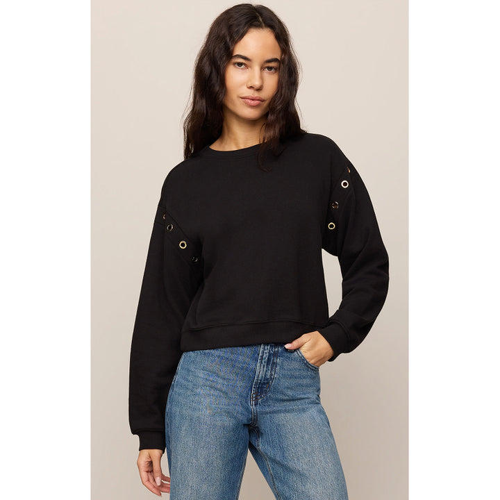 A woman modeling a black long-sleeve crewneck sweatshirt with gold grommet details on the sleeves, paired with blue jeans, against a solid light beige background.