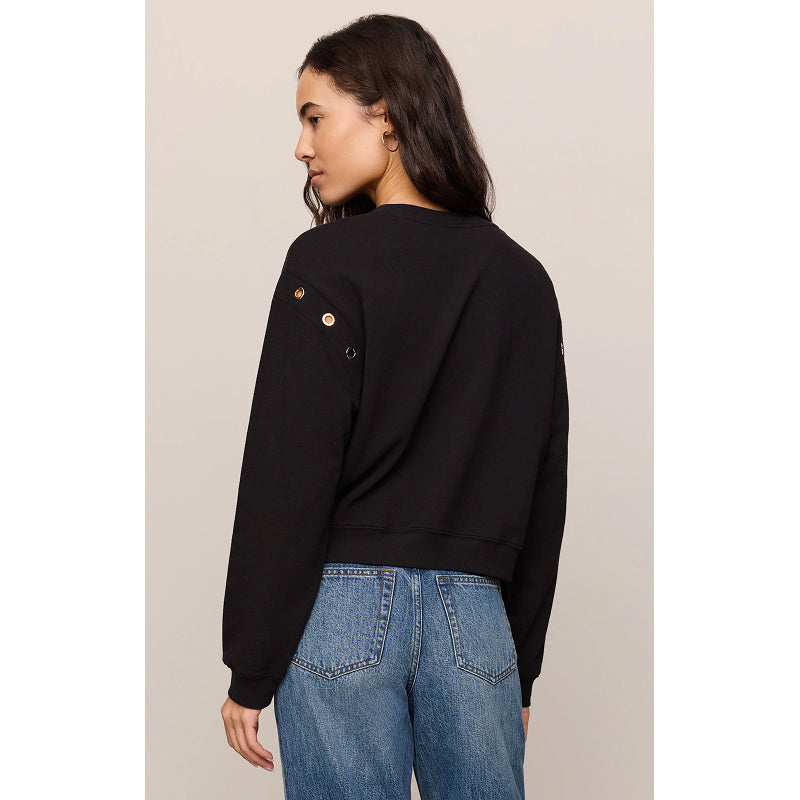 A woman shown from the back wearing a black cropped sweatshirt with decorative gold-tone grommets on the shoulder and blue jeans, against a plain light beige studio background.