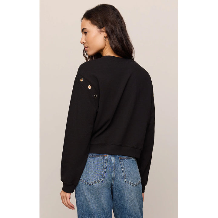A woman shown from the back wearing a black cropped sweatshirt with decorative gold-tone grommets on the shoulder and blue jeans, against a plain light beige studio background.