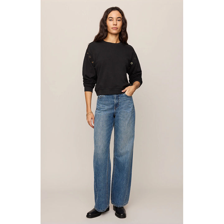 A woman modeling a black long-sleeve top with decorative grommets on the shoulders and wide-leg blue jeans, standing against a plain beige studio background.