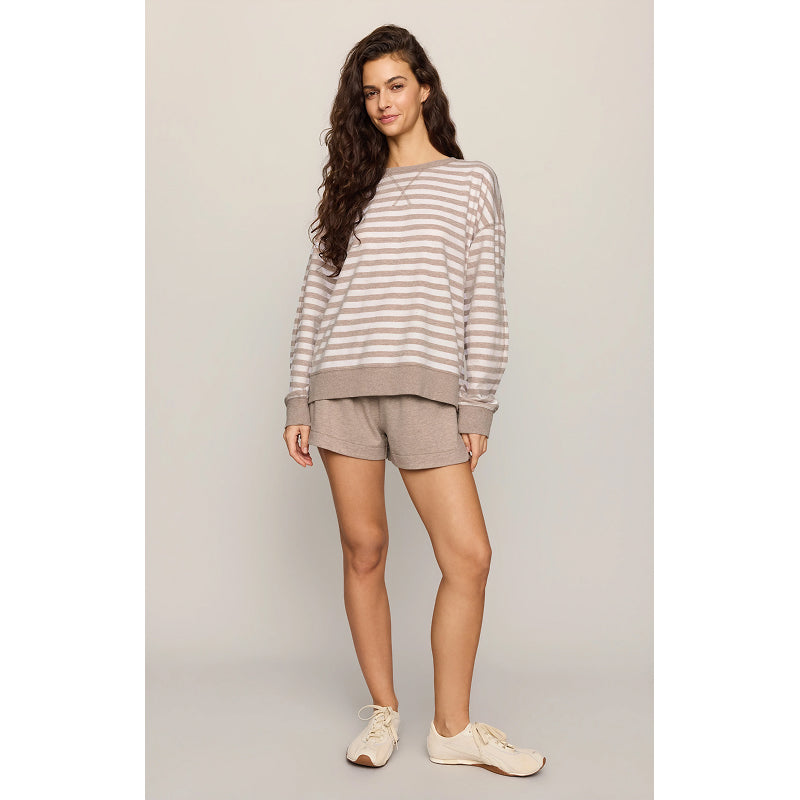 A woman modeling a matching lounge set consisting of a beige and taupe striped long-sleeve sweatshirt and solid taupe shorts, paired with cream sneakers, against a neutral studio backdrop.