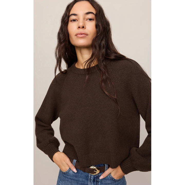 A woman wearing a dark brown knit crewneck sweater and blue jeans with a gold-belt, standing against a neutral beige background.