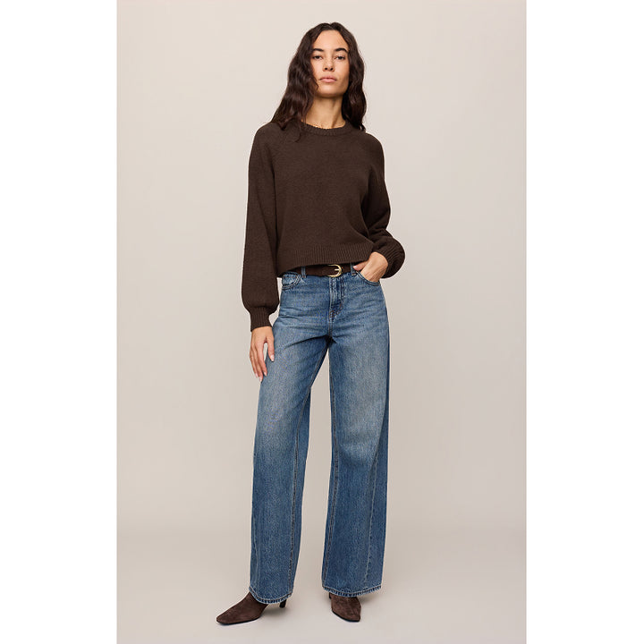 A woman modeling a dark brown long-sleeve knit sweater and wide-leg blue jeans against a plain beige studio background.
