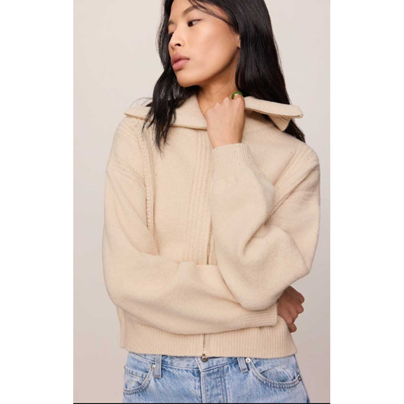 A woman wearing a cream-colored knit zip-up hoodie with a wide collar and ribbed hem, paired with light blue jeans, set against a plain light beige background.
