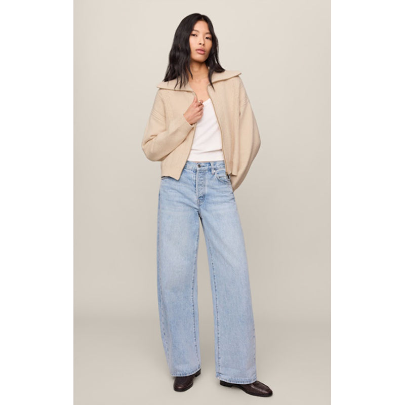 A model stands against a plain beige studio background wearing a light beige cropped jacket over a white top and light-wash wide-leg jeans.