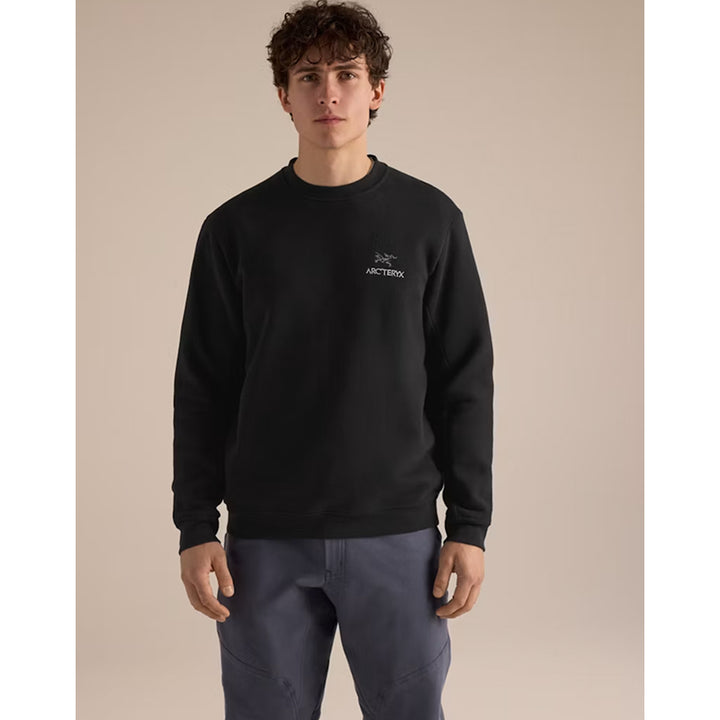A man wearing a black Arc'teryx crewneck sweatshirt and slate blue pants, standing against a solid beige studio background.