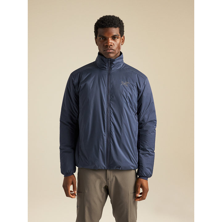 A man wearing a navy blue Arc'teryx puffer jacket and gray cargo pants, standing against a solid beige studio background.