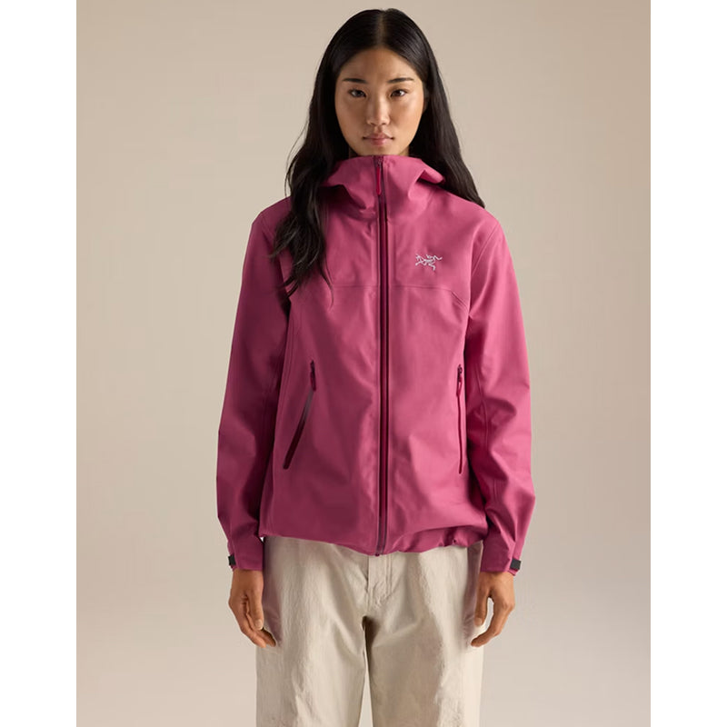 A woman modeling a pink Arc'teryx softshell jacket with a full-zip front, zippered hand pockets, and a small embroidered logo on the left chest, paired with light beige pants against a solid beige background.