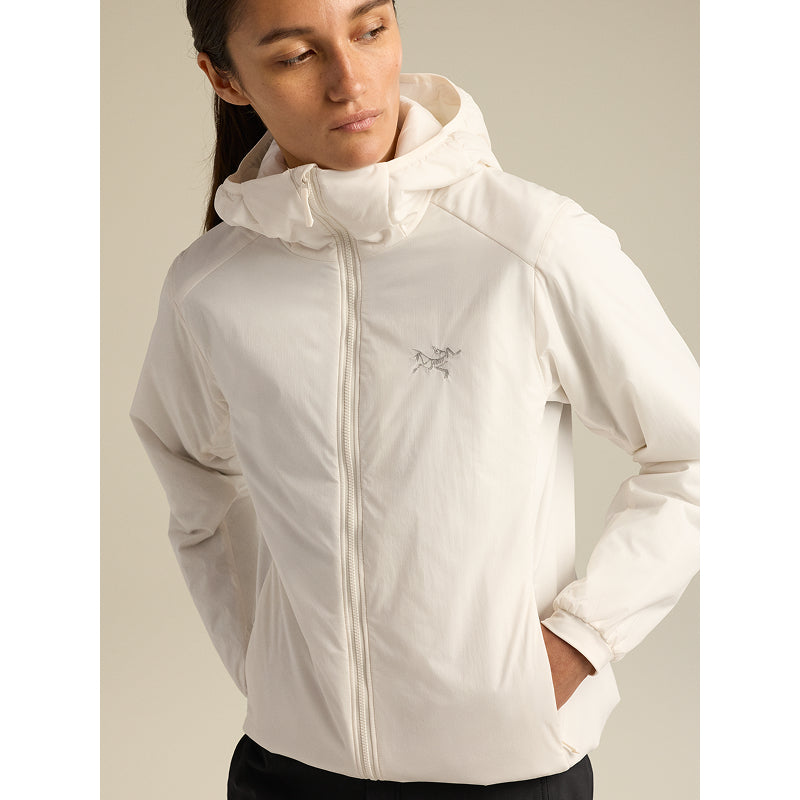 A woman wearing a light off-white hooded jacket with a small embroidered logo on the left chest, photographed against a plain beige studio background.
