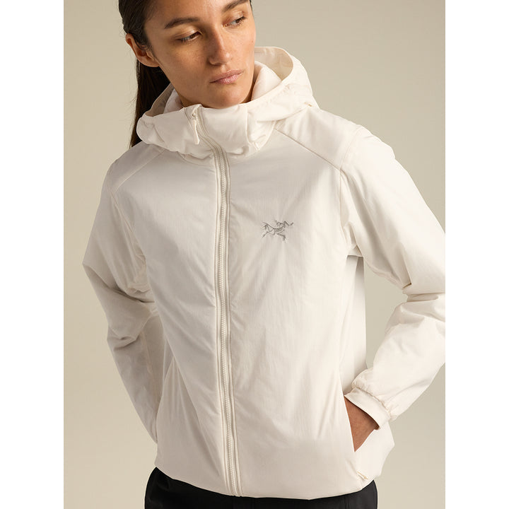 A woman wearing a light off-white hooded jacket with a small embroidered logo on the left chest, photographed against a plain beige studio background.