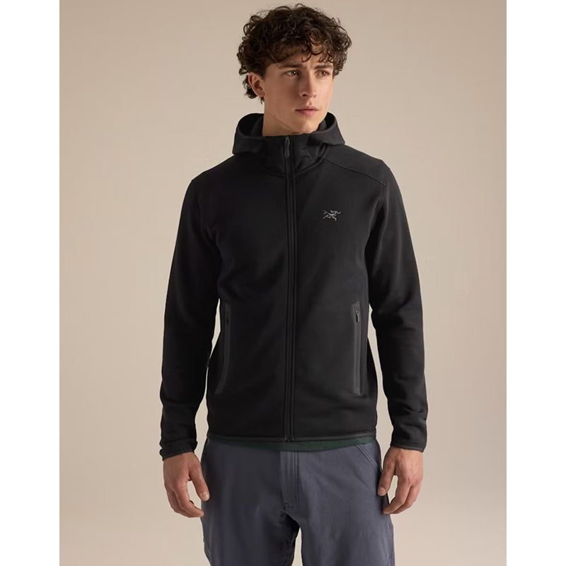 A man wearing a black Arc'teryx zip-up hoodie with a small white logo on the left chest, paired with slate blue pants, standing against a solid beige background.