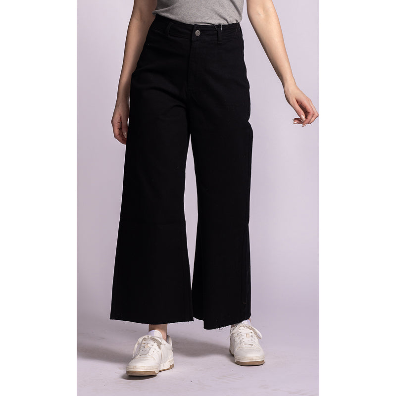A model wearing black wide-leg cropped pants, styled with a gray t-shirt and white sneakers, standing against a light lavender studio background.