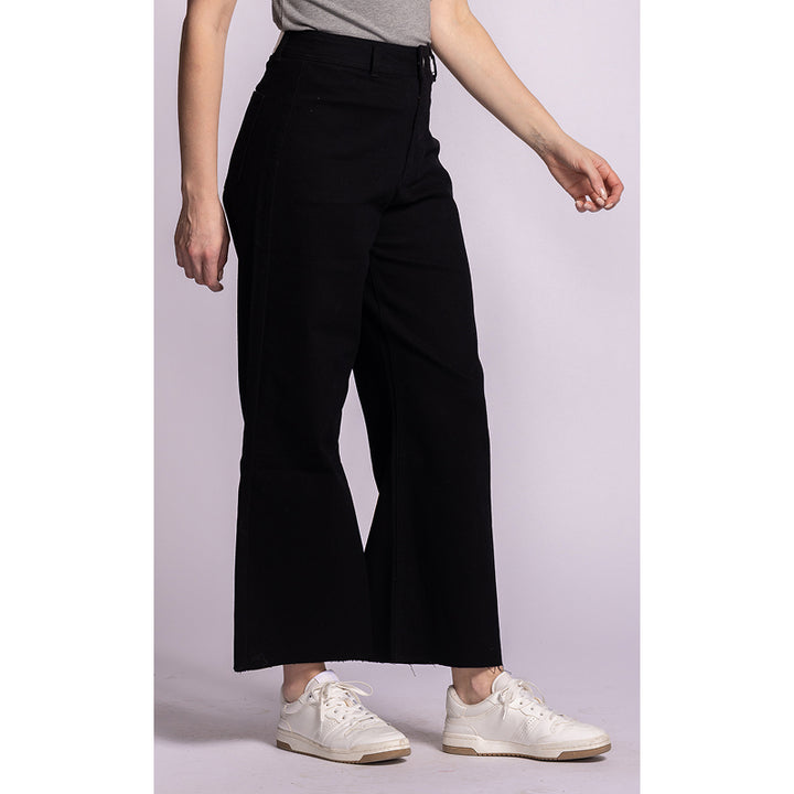 A model wearing black wide-leg cropped pants, a gray top, and white sneakers, shown from the waist down against a light lavender studio background.