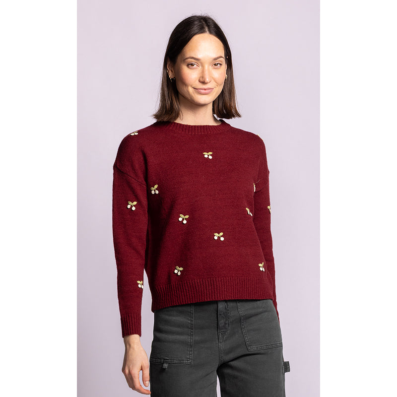 A woman modeling a burgundy knit sweater with small yellow embroidered floral motifs, paired with dark gray pants, against a solid light purple background.