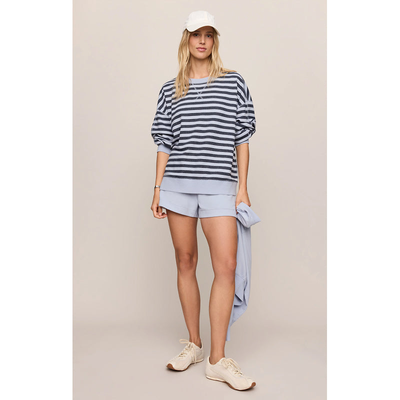 A woman modeling a navy and gray striped long-sleeve top, light blue shorts, a white baseball cap, and beige sneakers against a solid beige studio background.