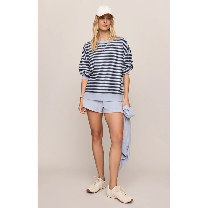 A woman modeling a navy and gray striped long-sleeve top, light blue shorts, a white baseball cap, and beige sneakers against a solid beige studio background.