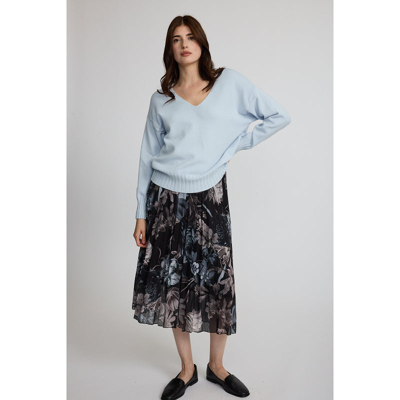 A woman modeling a light blue knit V-neck sweater paired with a dark floral midi skirt and black loafers, standing against a neutral light gray studio background.