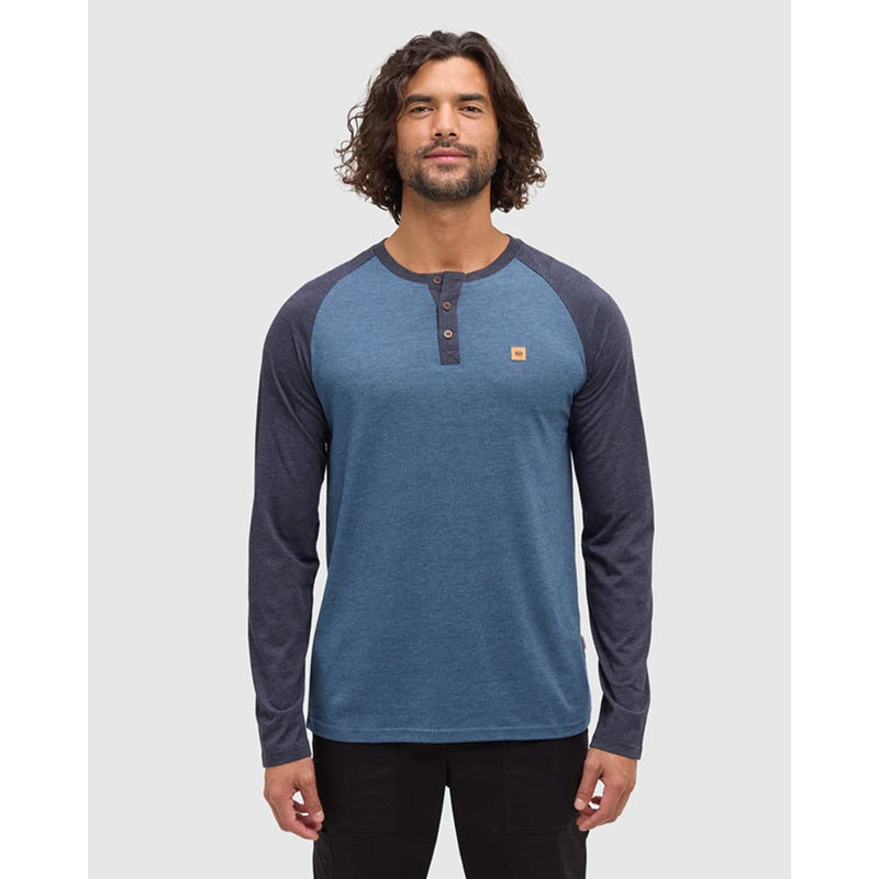 A man wearing a blue and navy raglan henley shirt with a small orange logo on the chest, standing against a light gray background.