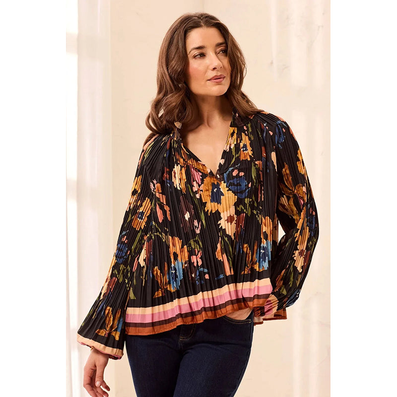 A woman wearing a black pleated long-sleeve blouse with a vibrant multicolor floral print and a pink, orange, and yellow striped hem, paired with dark blue jeans.