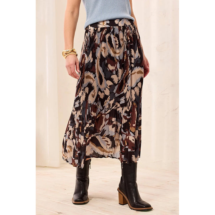 A woman models a high-waisted, flowy midi skirt with an abstract ikat-style pattern in brown, beige, and black. She is wearing a light blue ribbed top and black heeled ankle boots, standing against a neutral cream-colored curtain background.