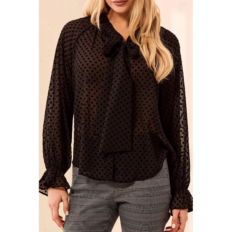 A woman wearing a black sheer long-sleeve blouse with a polka dot pattern and a large bow at the collar, paired with gray and white striped trousers.