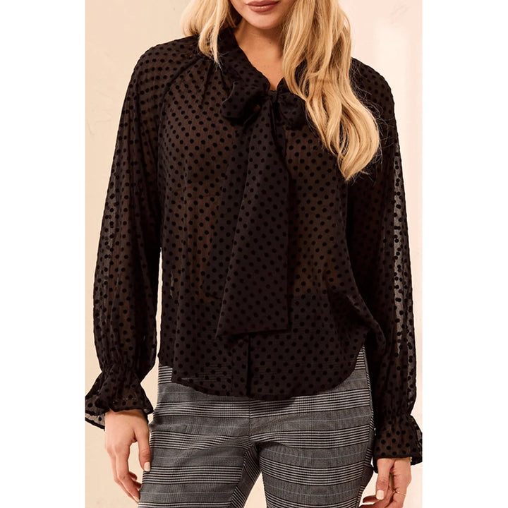 A woman wearing a black sheer long-sleeve blouse with a polka dot pattern and a large bow at the collar, paired with gray and white striped trousers.