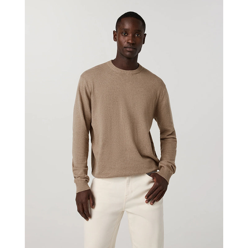A man modeling a light brown crewneck knit sweater paired with off-white trousers, standing against a neutral light gray background.