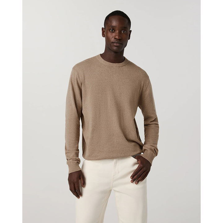 A man modeling a light brown crewneck knit sweater paired with off-white trousers, standing against a neutral light gray background.