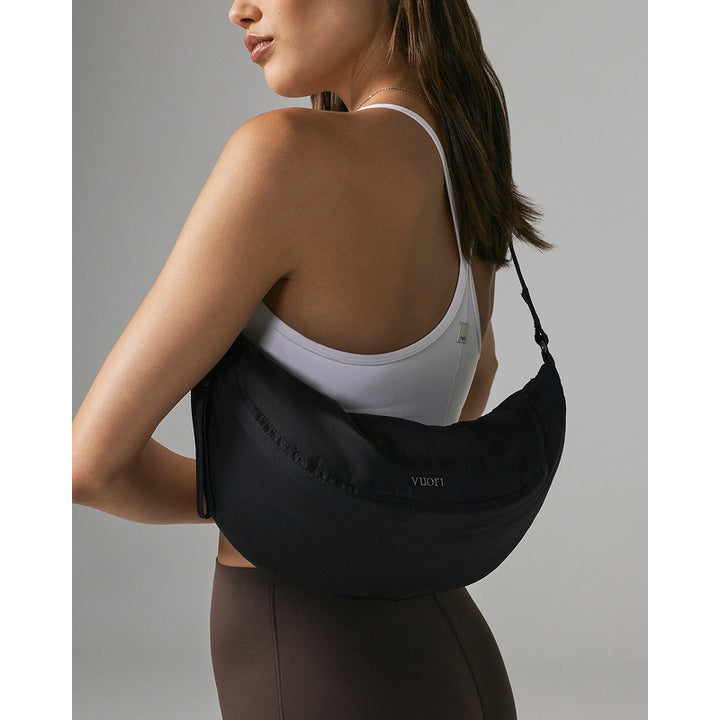 A woman wearing a white sports bra and brown leggings carries a black crescent-shaped shoulder bag with a thin strap. The bag has a small white 'vuori' logo on the front, set against a solid gray studio background.