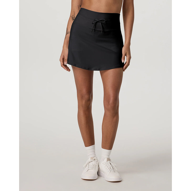 A model wearing a black athletic mini skirt with a drawstring waist, paired with white crew socks and white sneakers, shown from the waist down against a light gray studio background.