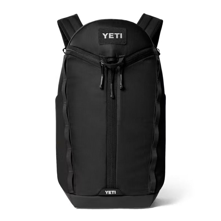 A black YETI backpack shown from the front on a clean white background. The backpack features a top zip closure, a central vertical zipper, side compression straps, and the YETI brand name visible on the top and bottom patches.