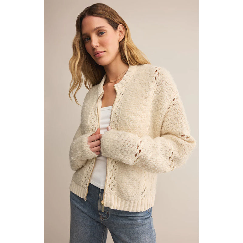 Woman wearing a beige knitted cardigan over a white shirt and blue jeans on a plain background