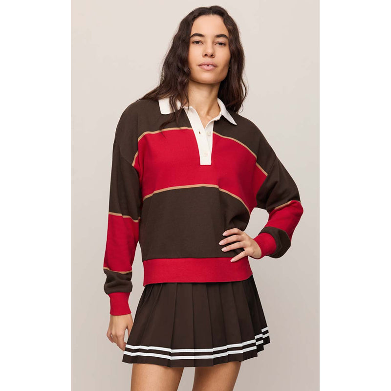 A woman modeling a color-blocked long-sleeve rugby-style top and pleated mini skirt set. The set features dark brown, red, and white color blocking with a white collar and button placket on the top, and white stripes on the hem of the skirt.
