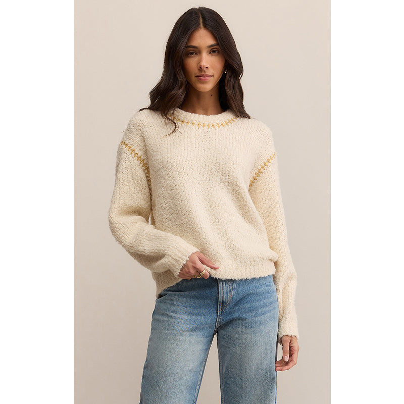 Woman wearing a cream sweater with gold accents and blue jeans on a beige background