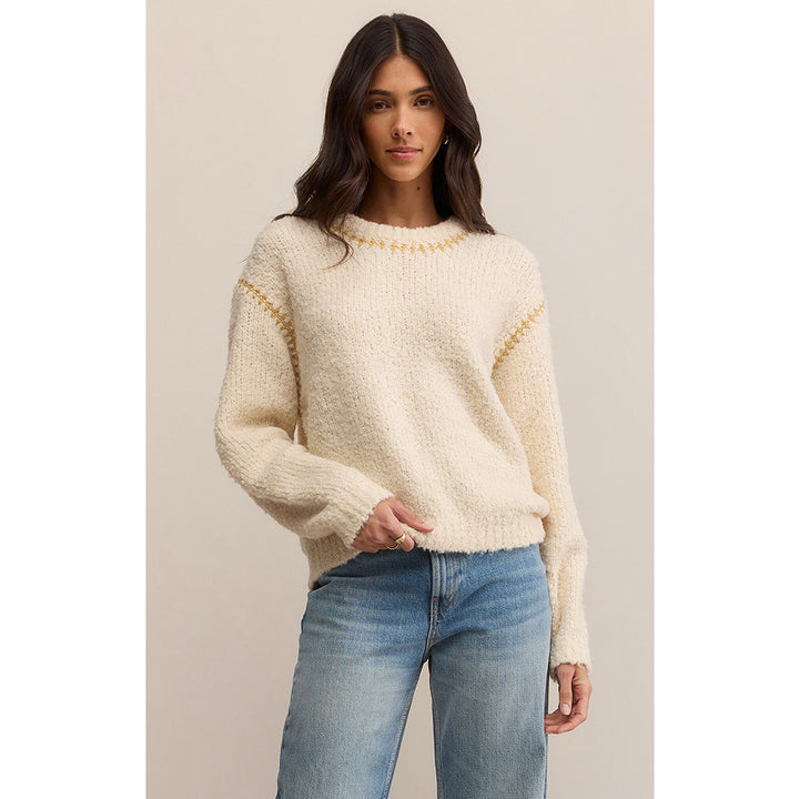 Woman wearing a cream sweater with gold accents and blue jeans on a beige background